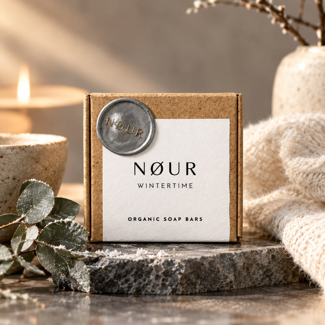 NØUR Wintertime Naturseife in Graspapier-Schachtel mit Siegel, auf Steinplatte zwischen Eukalyptuszweigen, Kerze und Strickdecke