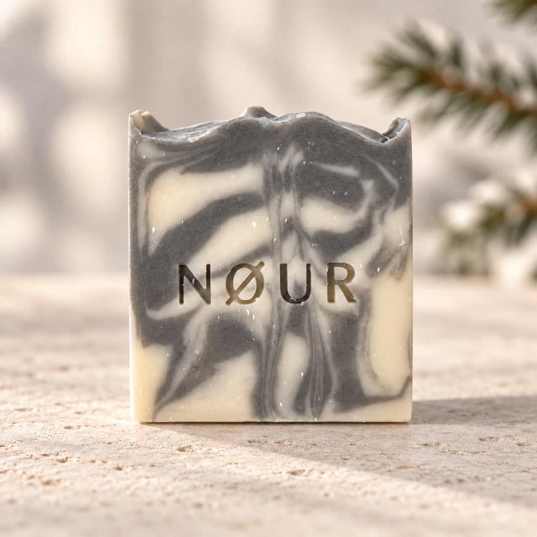 NØUR Naturseife Wintertime mit grau-weißer Marmorierung und geprägtem Logo auf hellem Stein, im Hintergrund ein Tannenzweig